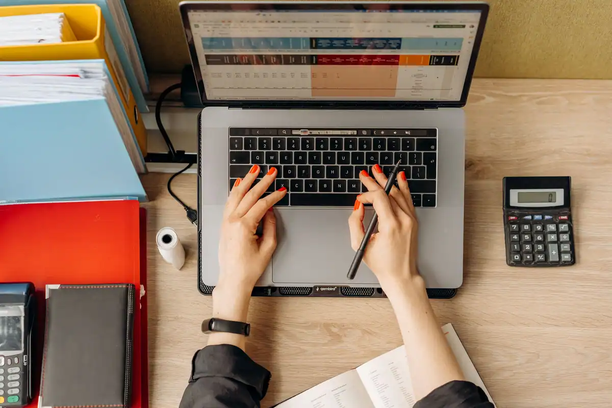 Mâini care tastează la laptop, văzute de sus, lângă dosare și un calculator pe birou