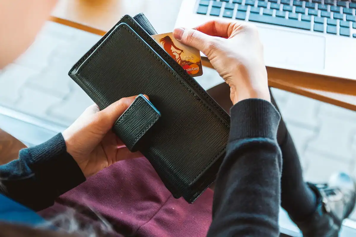 Mâini care scot un card dintr-un portofel negru, lângă un laptop