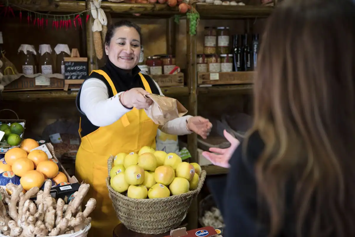 Vânzătoare zâmbitoare care înmânează o pungă de hârtie unui client, într-o alimentară