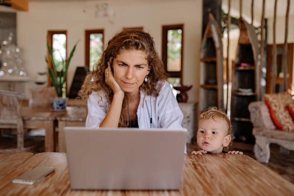 Femeie încruntată în fața laptopului, cu un copil mic lângă ea