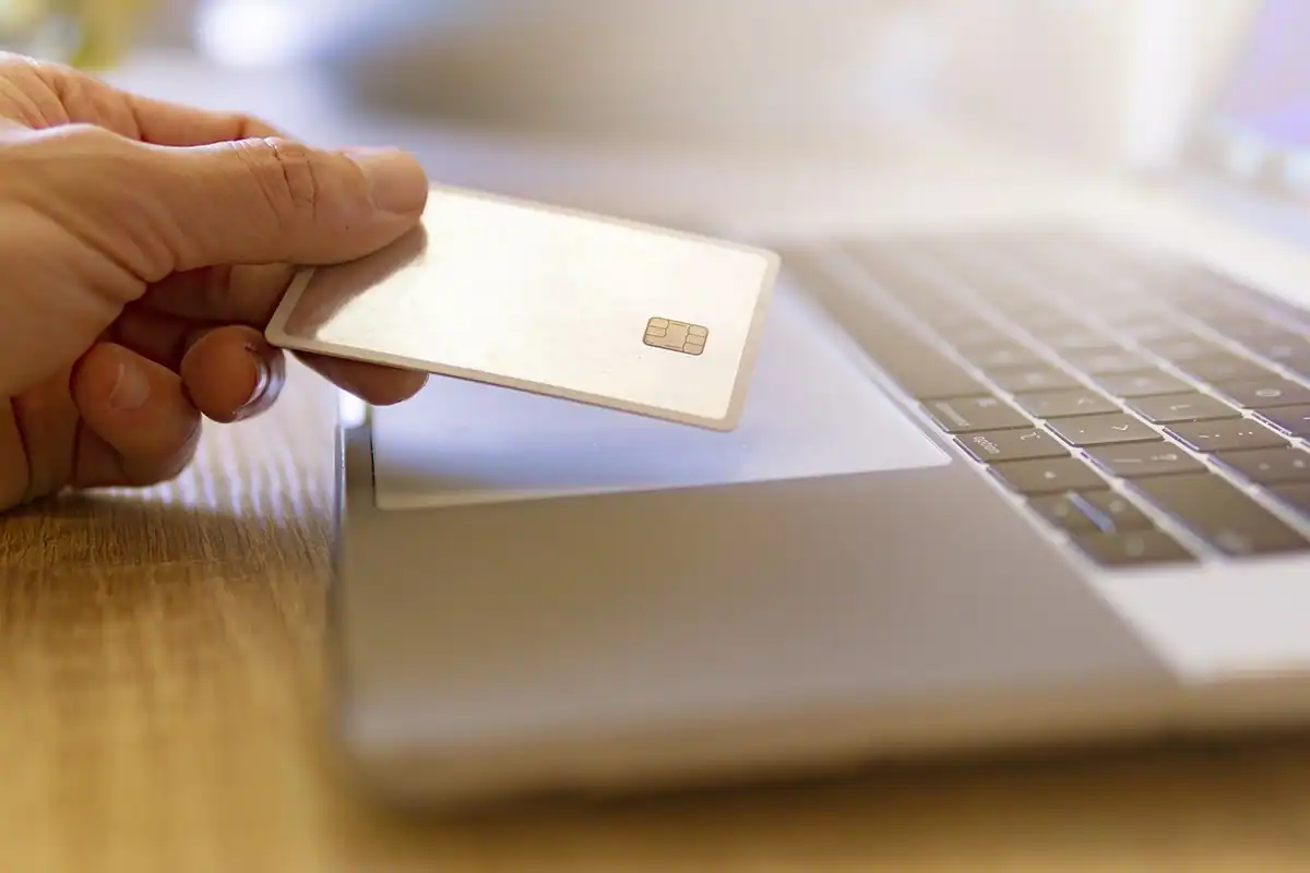 Mână care ține un card bancar deasupra unui laptop, în timpul unei plăți online