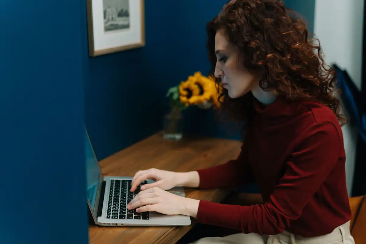 Femeie în pulover bordo tastează la laptop, la un birou cu floarea-soarelui