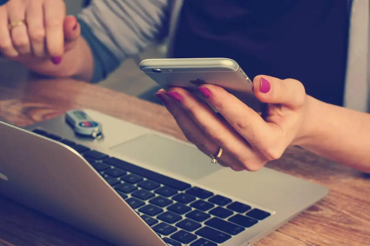 Mâini care țin un telefon mobil deasupra unui laptop, pe o masă de lemn